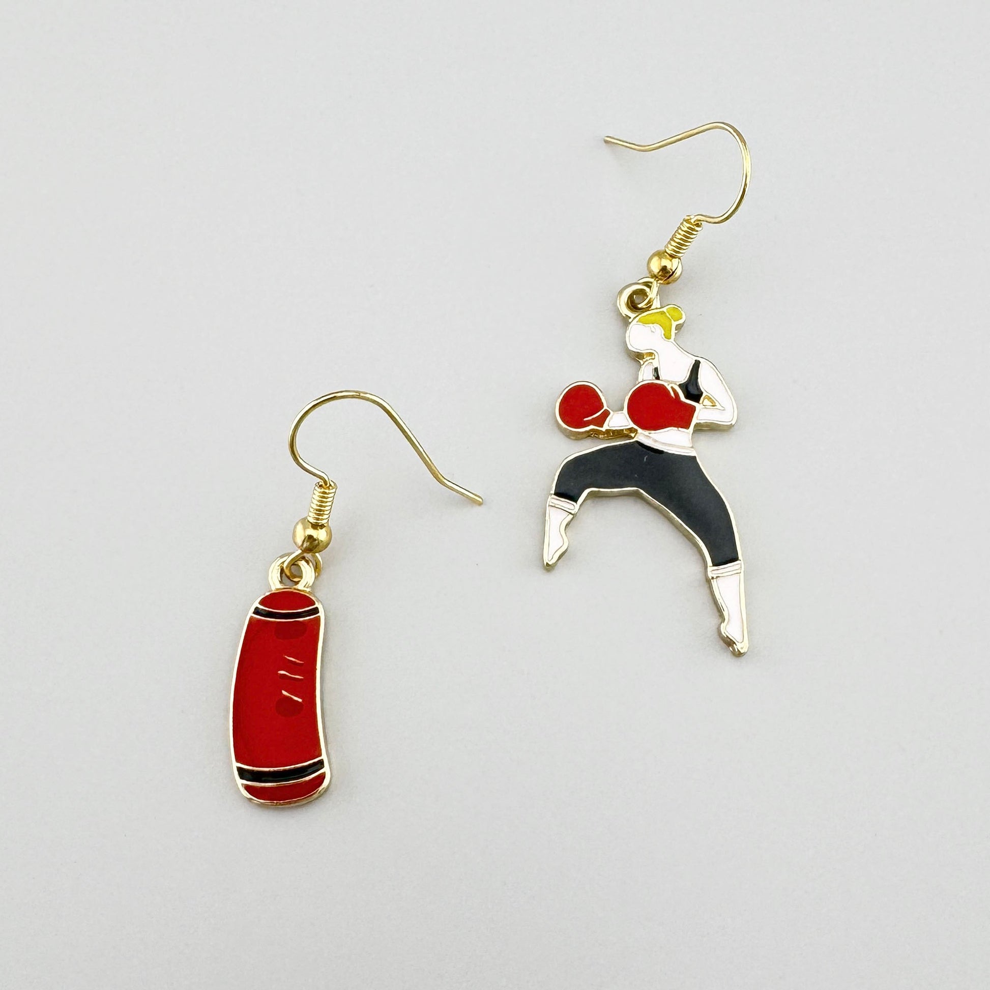 Kolczyki z motywem bokserskim w kolorze złotym z czerwoną emalią, przedstawiające bokserkę i worek treningowy.