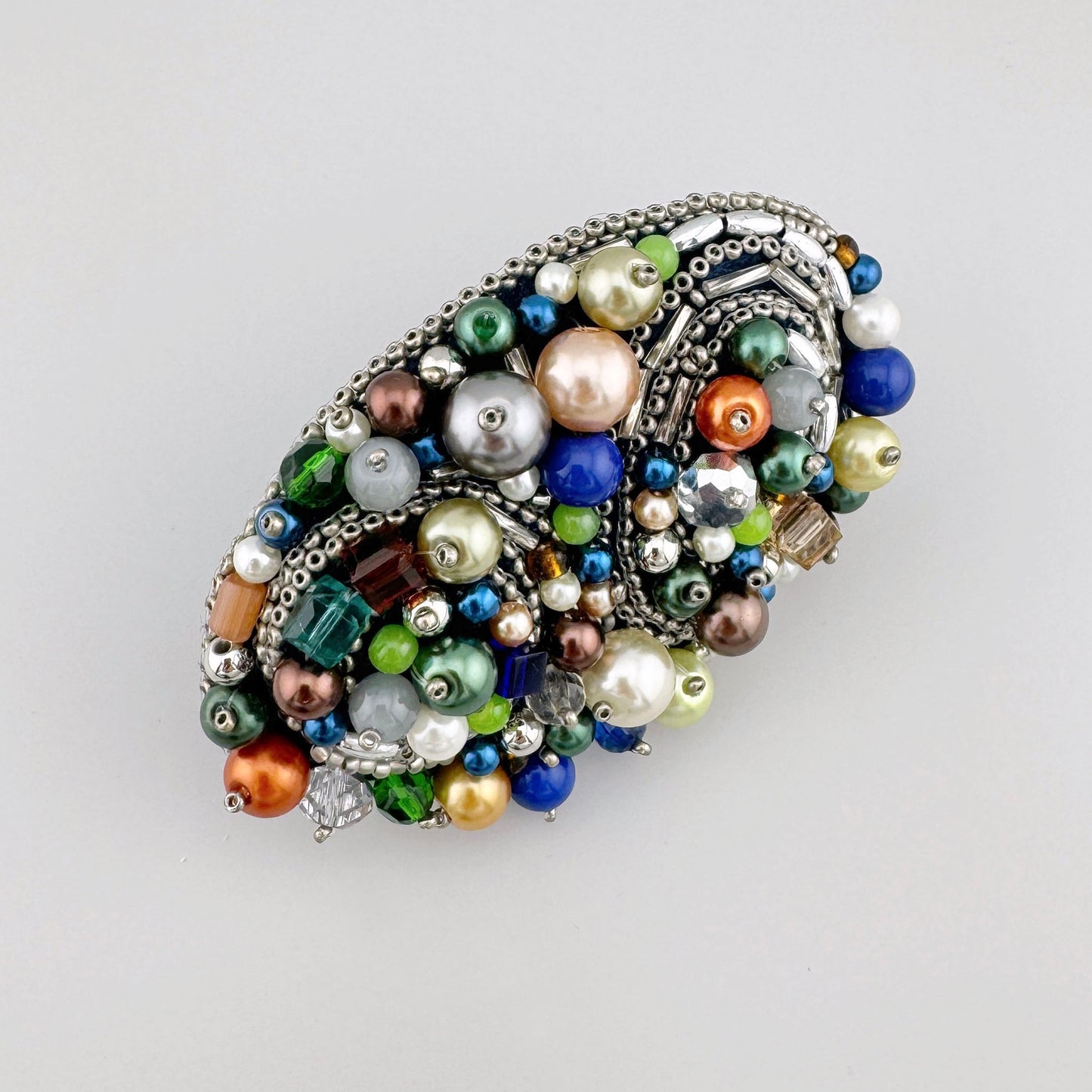 Broche ovale faite à la main, ornée de perles multicolores dans des tons de vert, bleu, or et argent.